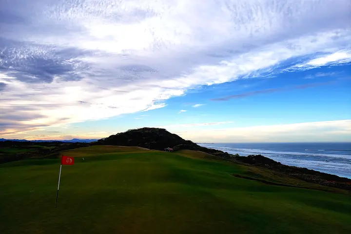 A green below a gorse-covered headland with the Pacific and coast range beyond.